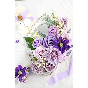 Purple artificial flower arrangement with roses and dahlias on a decorative mirror, with ribbon and small floral accents.