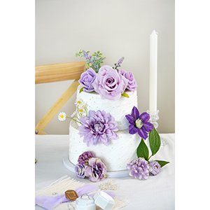 Two-tier white cake decorated with purple artificial flowers and greenery, with a white candle on the side.