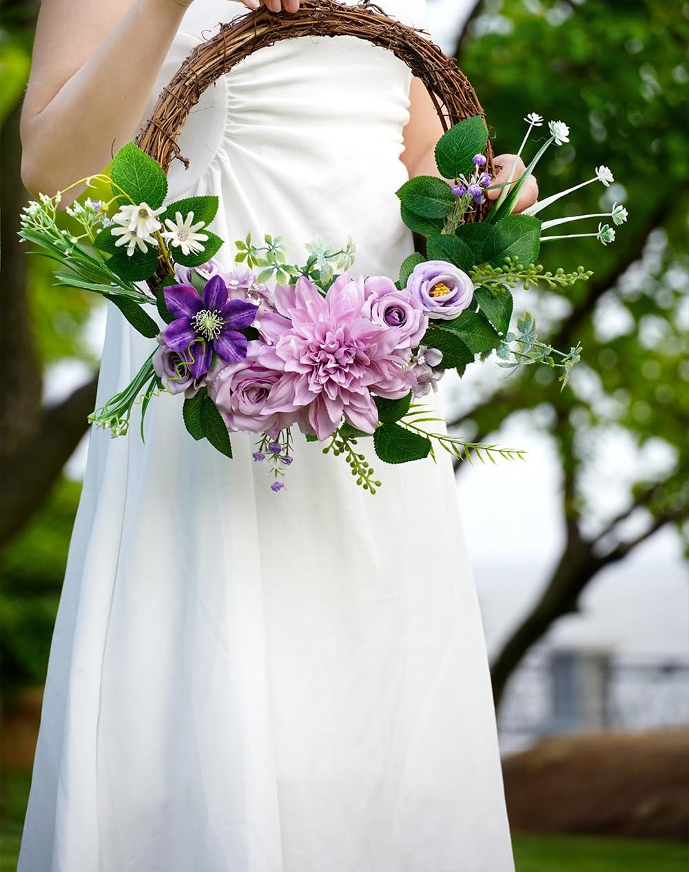 Woman in white dress holding a floral wreath with purple and pink artificial flowers and green leaves