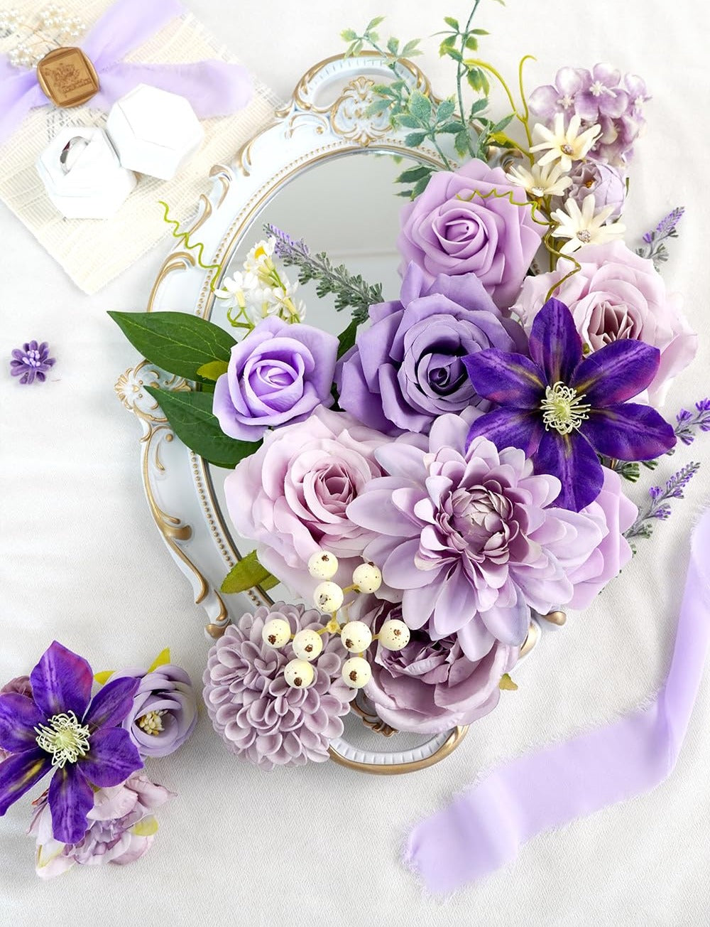 Assortment of purple artificial flowers arranged on a decorative mirror tray with wedding rings and ribbon.