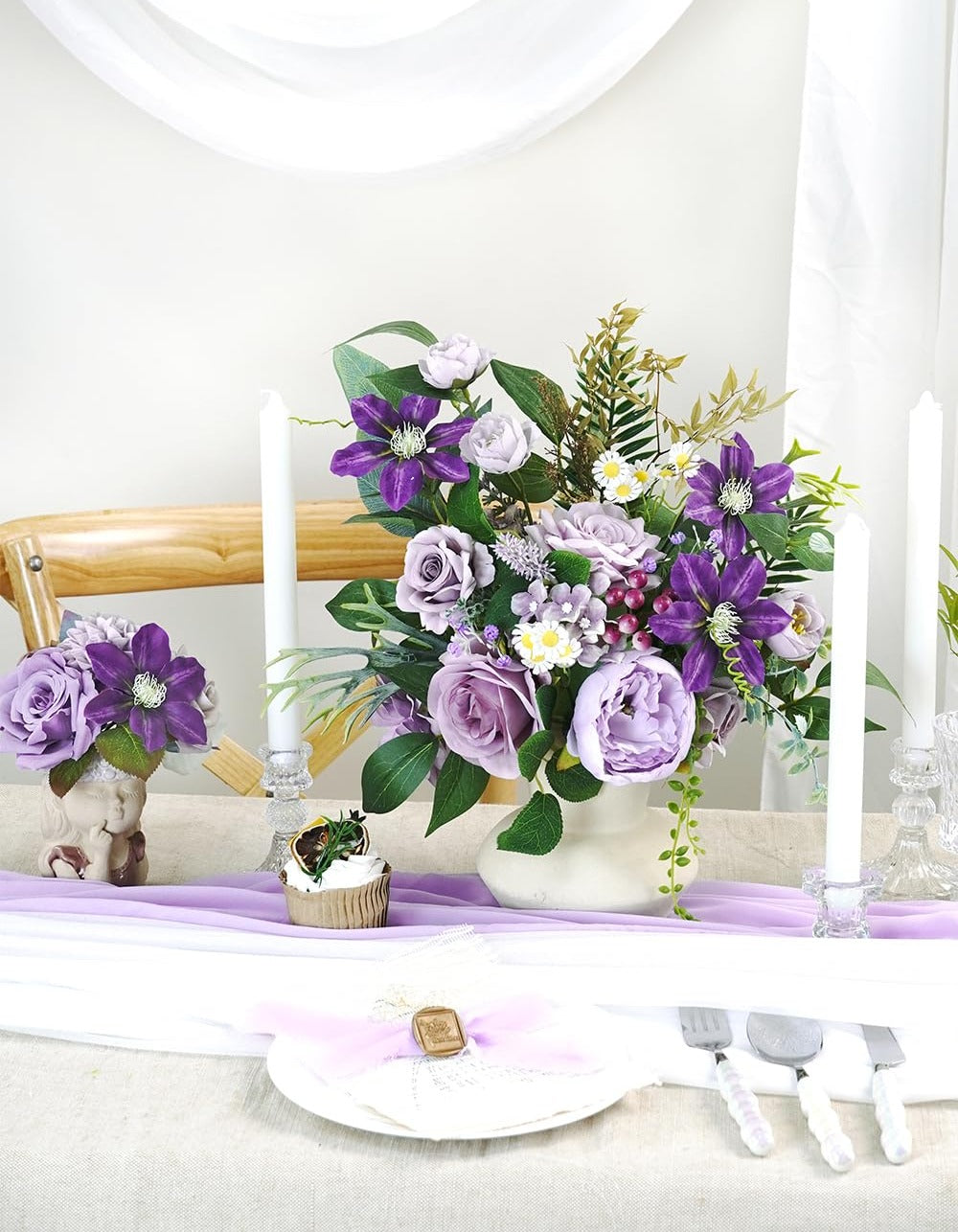 Purple artificial flower arrangement in a vase on a table with candles and place setting