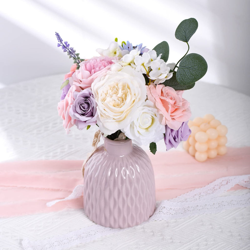 A pastel pink vase filled with a bouquet of artificial flowers including roses, lavender, and eucalyptus leaves, set on a white lace tablecloth.