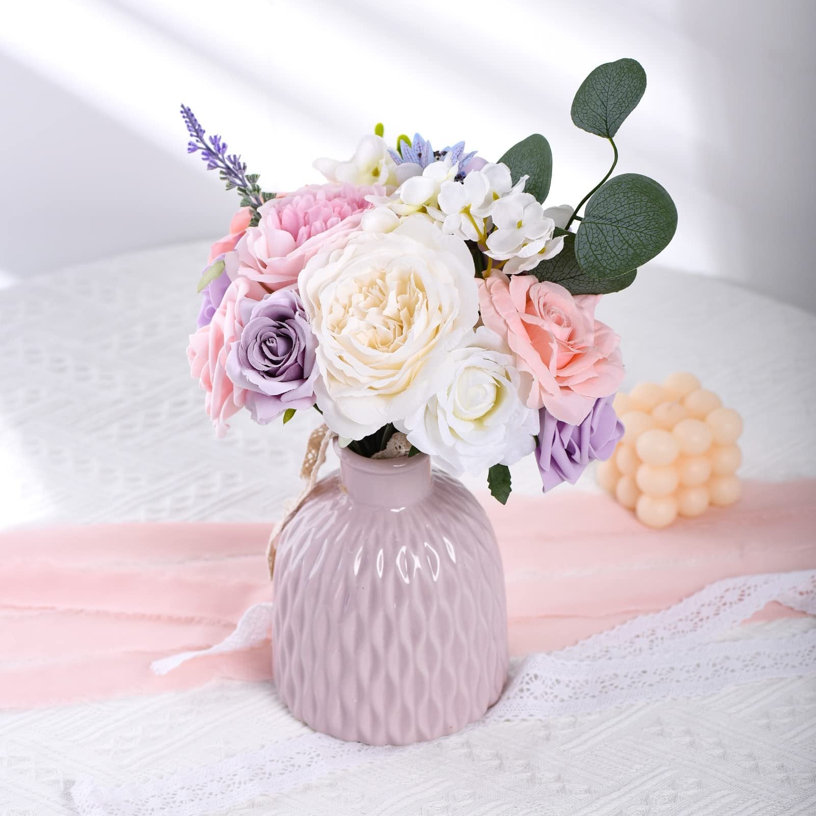 A pastel pink vase filled with a bouquet of artificial flowers including roses, lavender, and eucalyptus leaves, set on a white lace tablecloth.