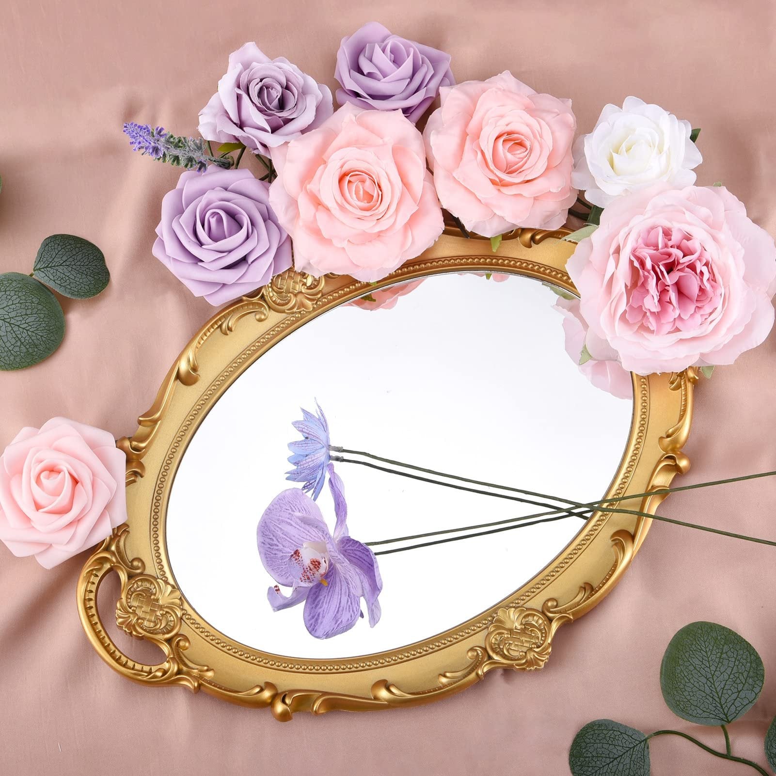 Assortment of artificial silk flowers in pink, purple, and white arranged around a gold ornate mirror on a pink satin background.