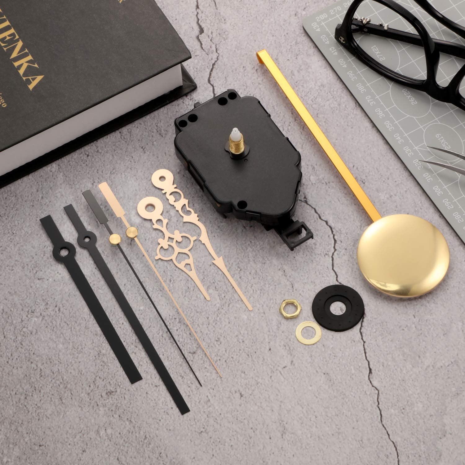 Quartz pendulum clock movement kit with hands and pendulum, laid out on a textured surface next to a book and glasses.