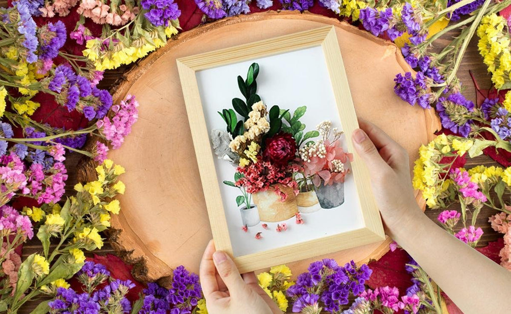 Hands holding a framed arrangement of dried flowers and leaves, surrounded by colorful dried flowers.