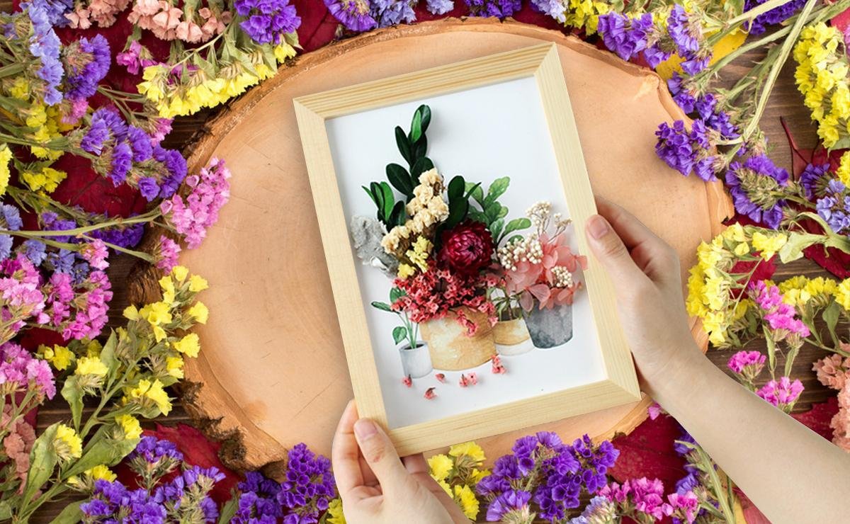 Hands holding a framed arrangement of dried flowers and leaves, surrounded by colorful dried flowers.