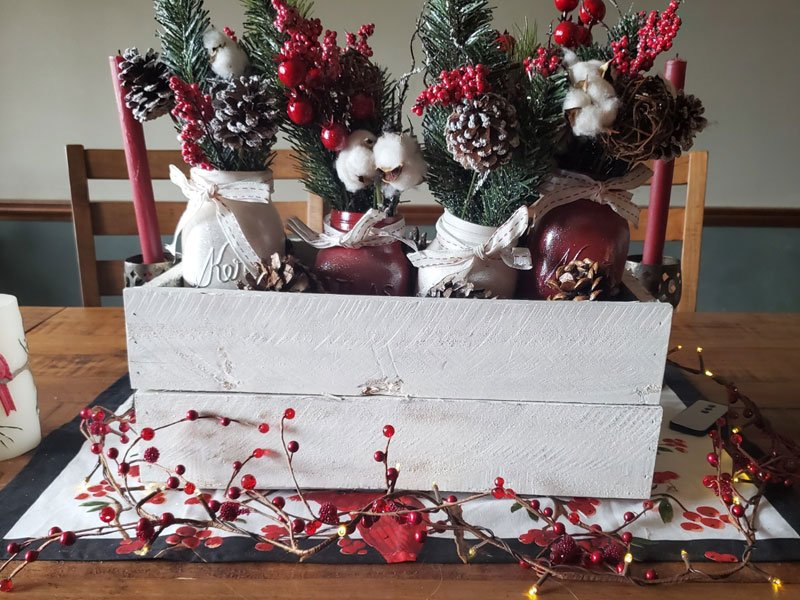 Christmas centerpiece with red berries, pinecones, and cotton in jars inside a white wooden crate.