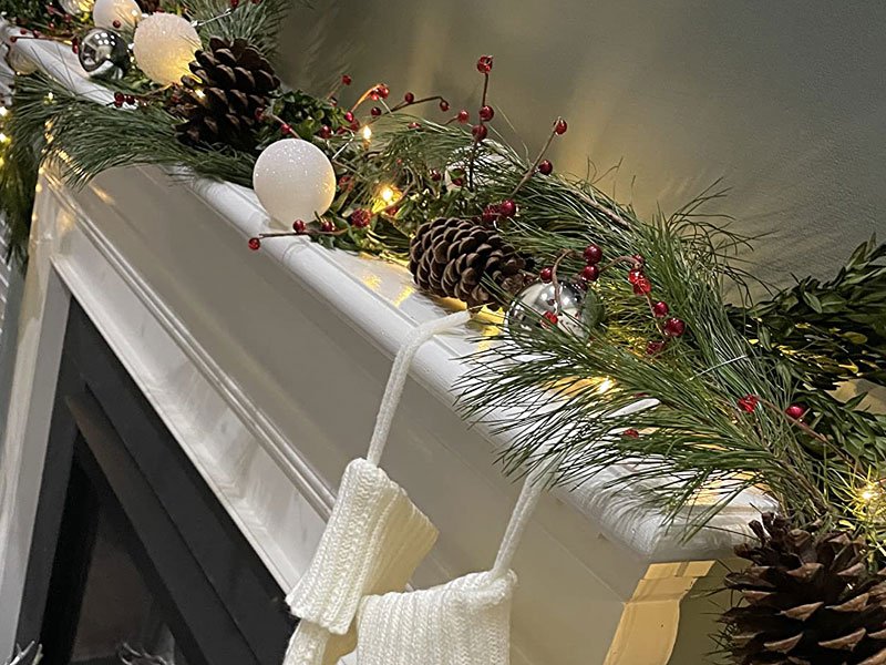 Pre-lit Christmas garland with pinecones, red berries, and warm white LED lights draped over a mantelpiece with white stockings.