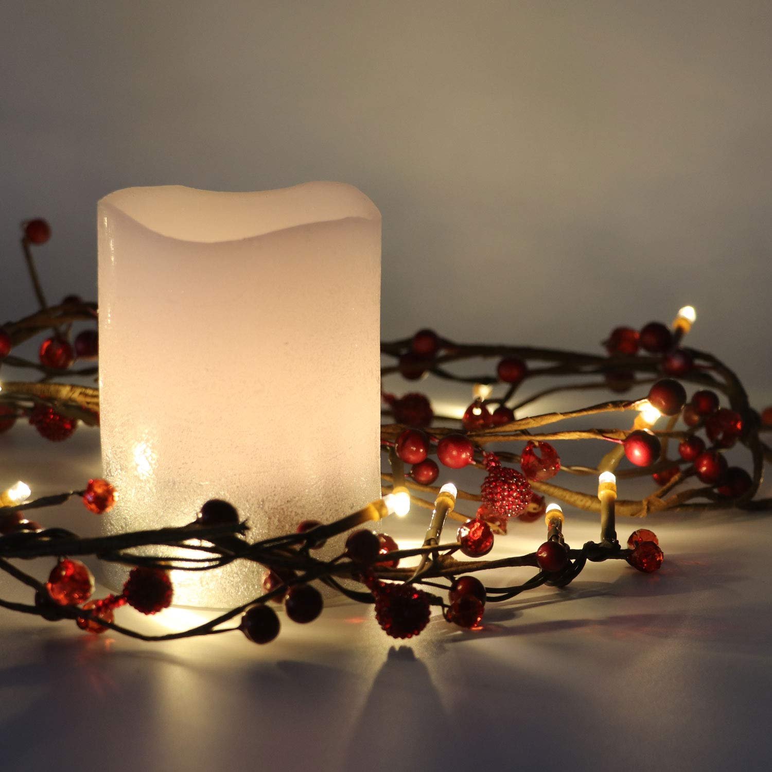 Red berry garland with warm white LED lights wrapped around a frosted white candle on a white surface.