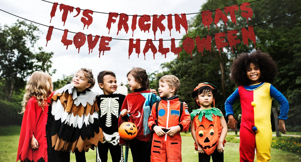 Group of children in Halloween costumes posing outdoors with a red glitter banner that says "It's Frickin Bats I Love Halloween"