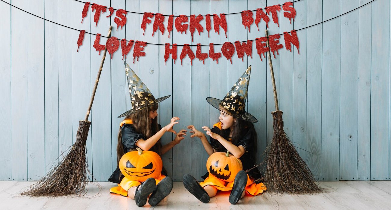 Two children in witch costumes sit with pumpkins and brooms, under a red glitter banner that reads 'It's Frickin Bats I Love Halloween'.