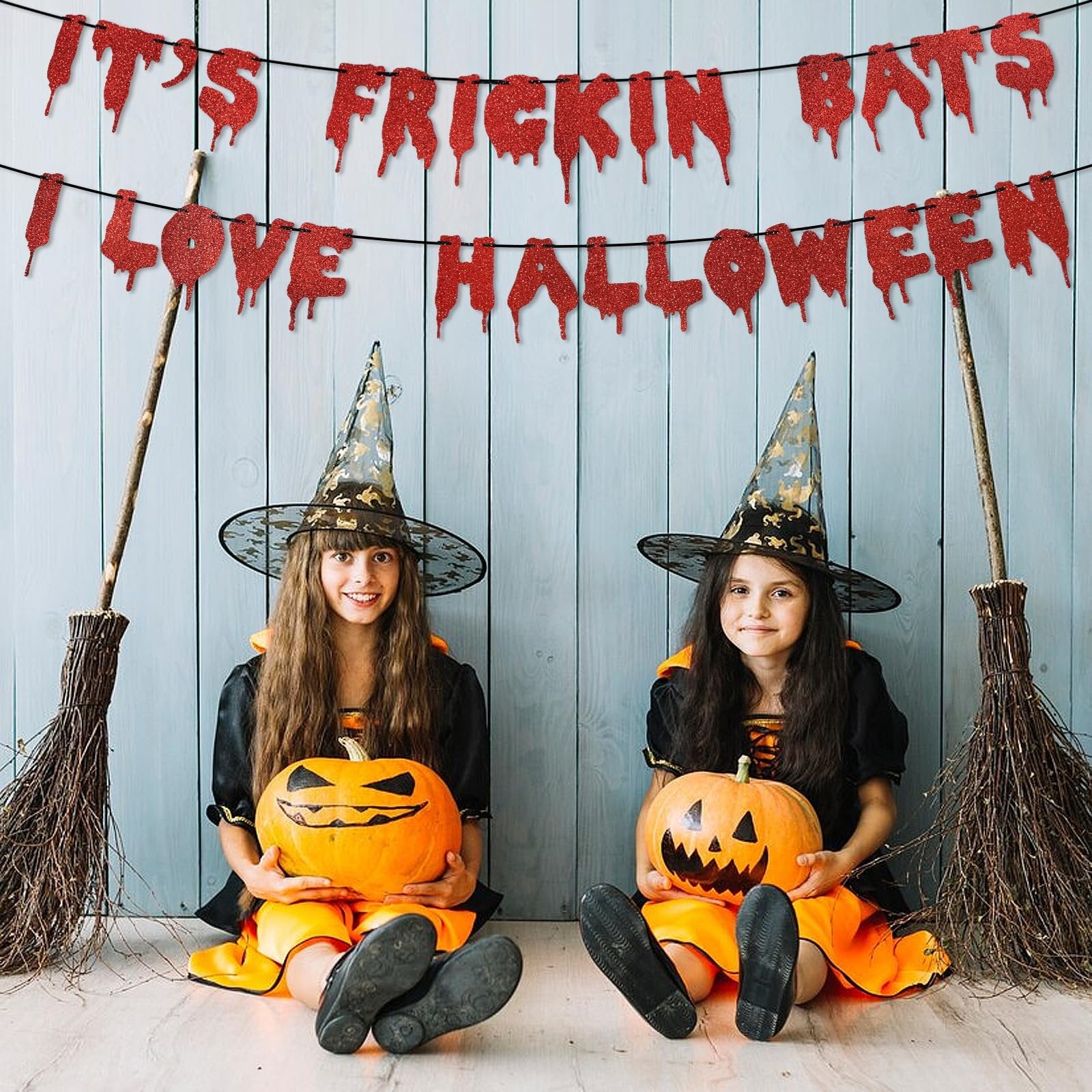 Two girls in witch costumes holding pumpkins, with a red glitter bat banner reading 'It's Frickin Bats I Love Halloween' behind them.