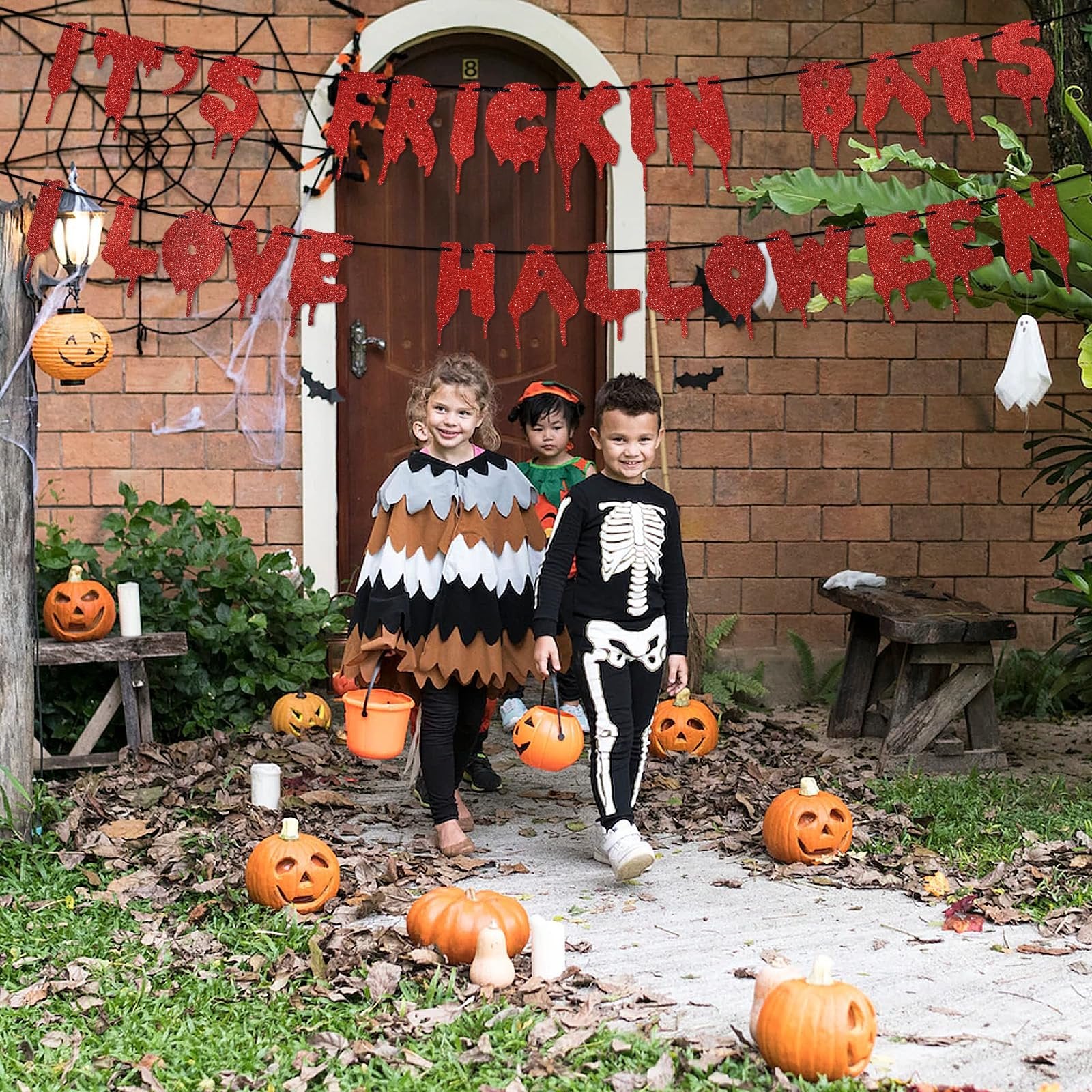 Three children in Halloween costumes walk down a path decorated with pumpkins and a glitter banner that reads "It's Frickin Bats I Love Halloween."
