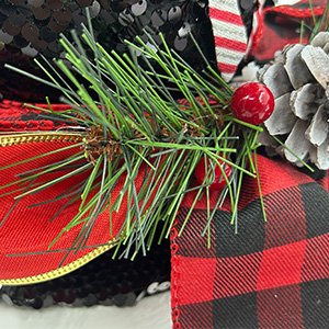 Close-up of a red velvet candy cane Christmas tree topper hat with sequins, pine sprigs, red berries, pinecone, and plaid ribbon.