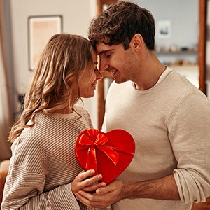 Couple holding a red heart-shaped gift box with a gold ribbon, indoors.