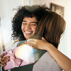 Two smiling women hugging, one holding a pink gift box with a ribbon.