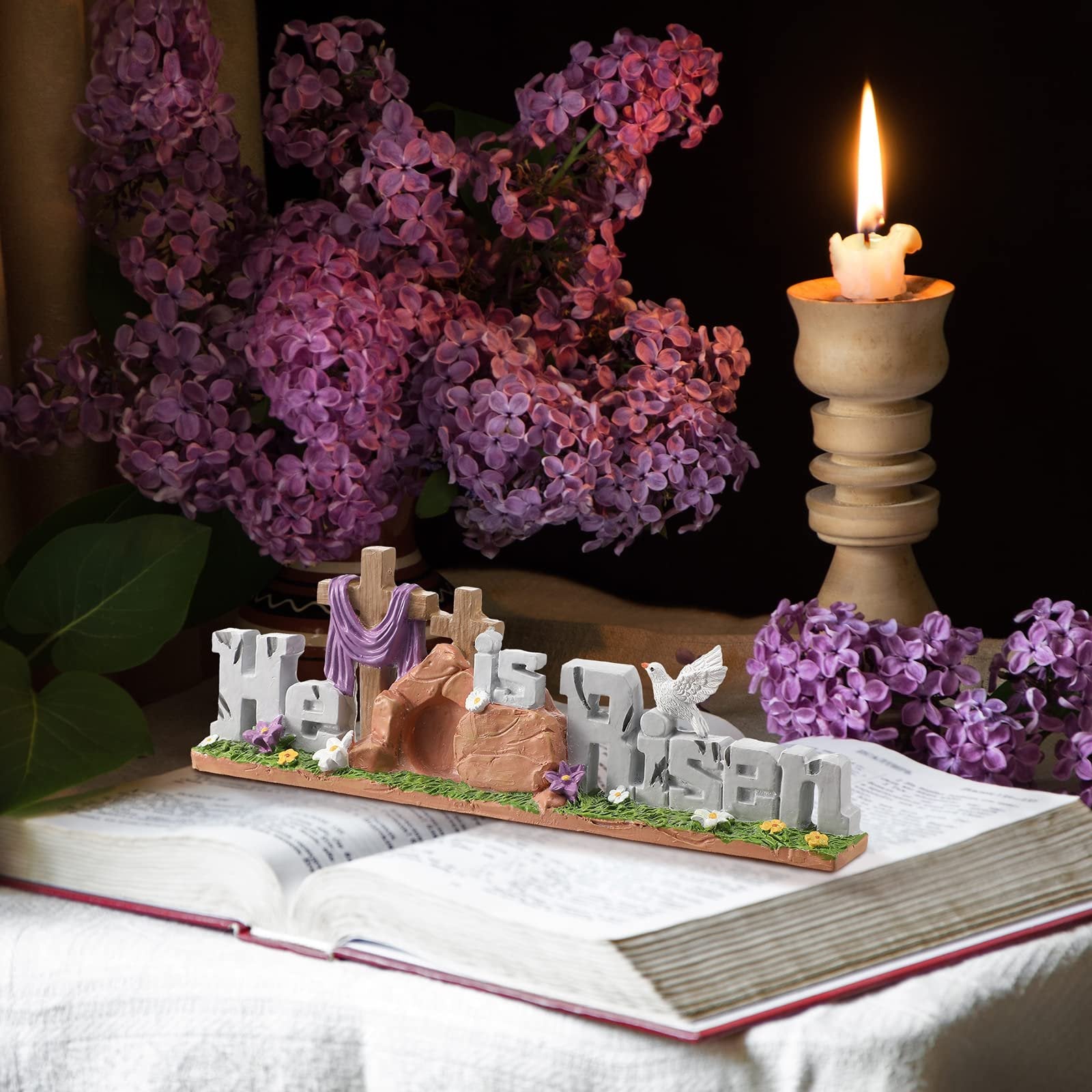 Easter figurine with 'He is Risen' text, crosses, tomb, and dove on an open book with lilacs and a candle.