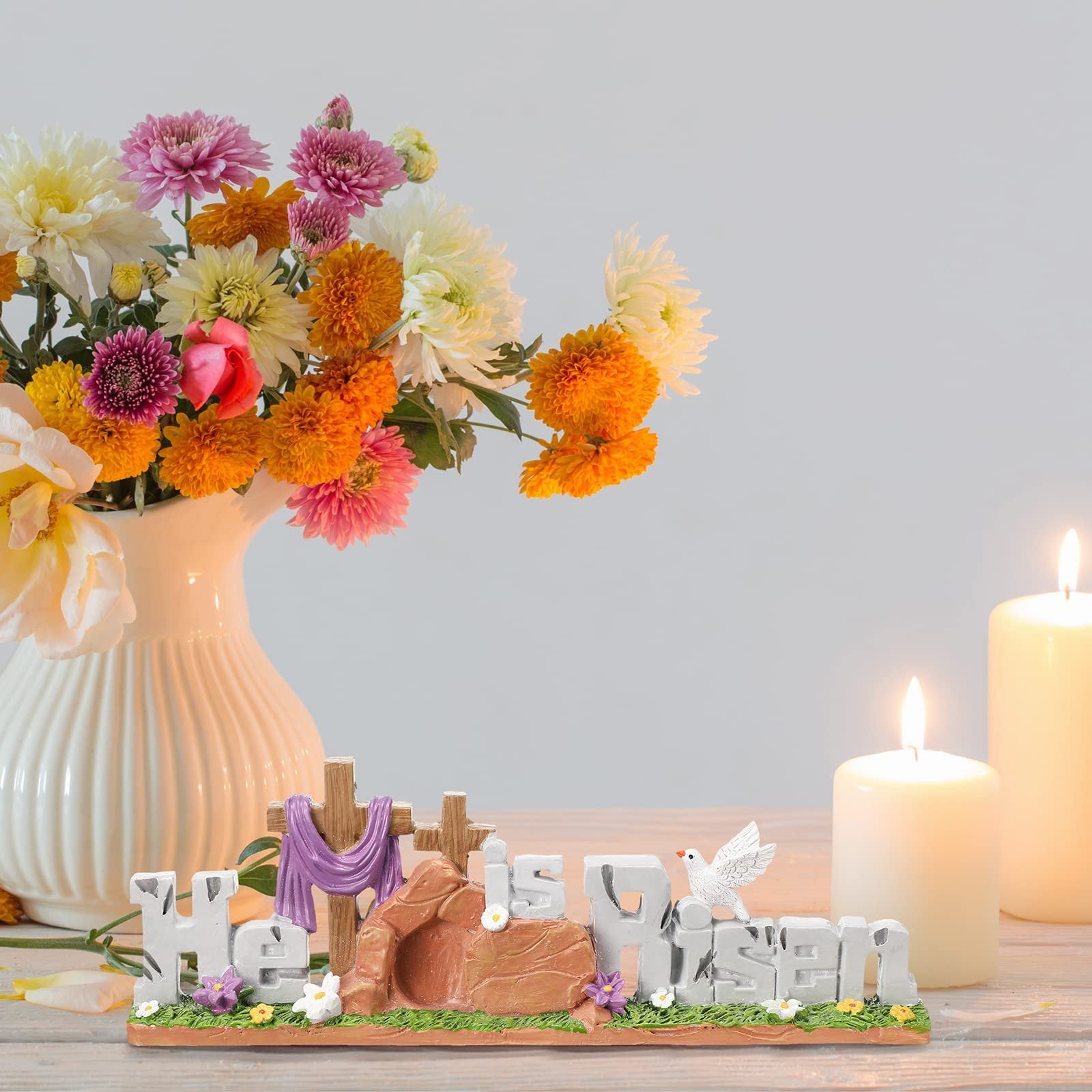 Easter figurine depicting the resurrection scene with crosses, tomb, dove, and text 'He is Risen', next to a vase of flowers and lit candles.