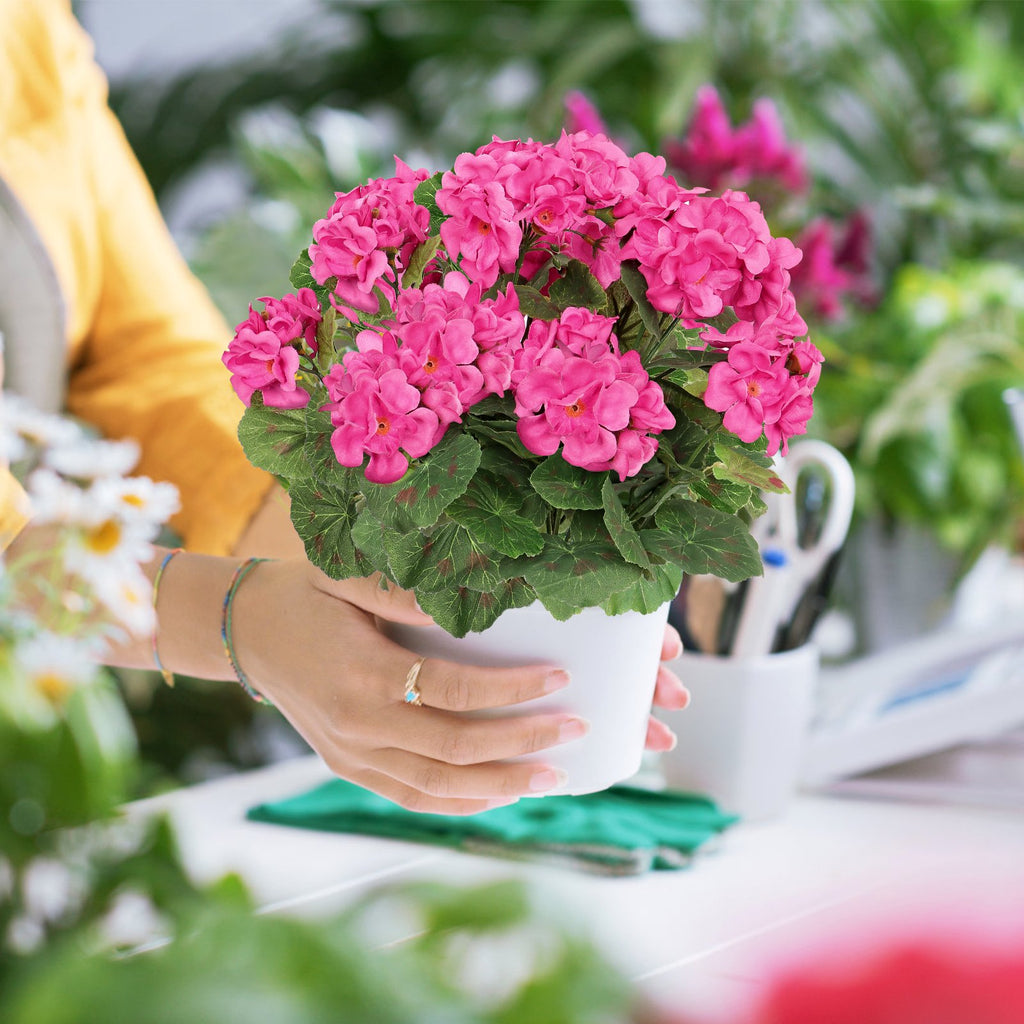 Hands holding a white pot with pink artificial geranium flowers and green leaves, with a blurred background of plants and office supplies.