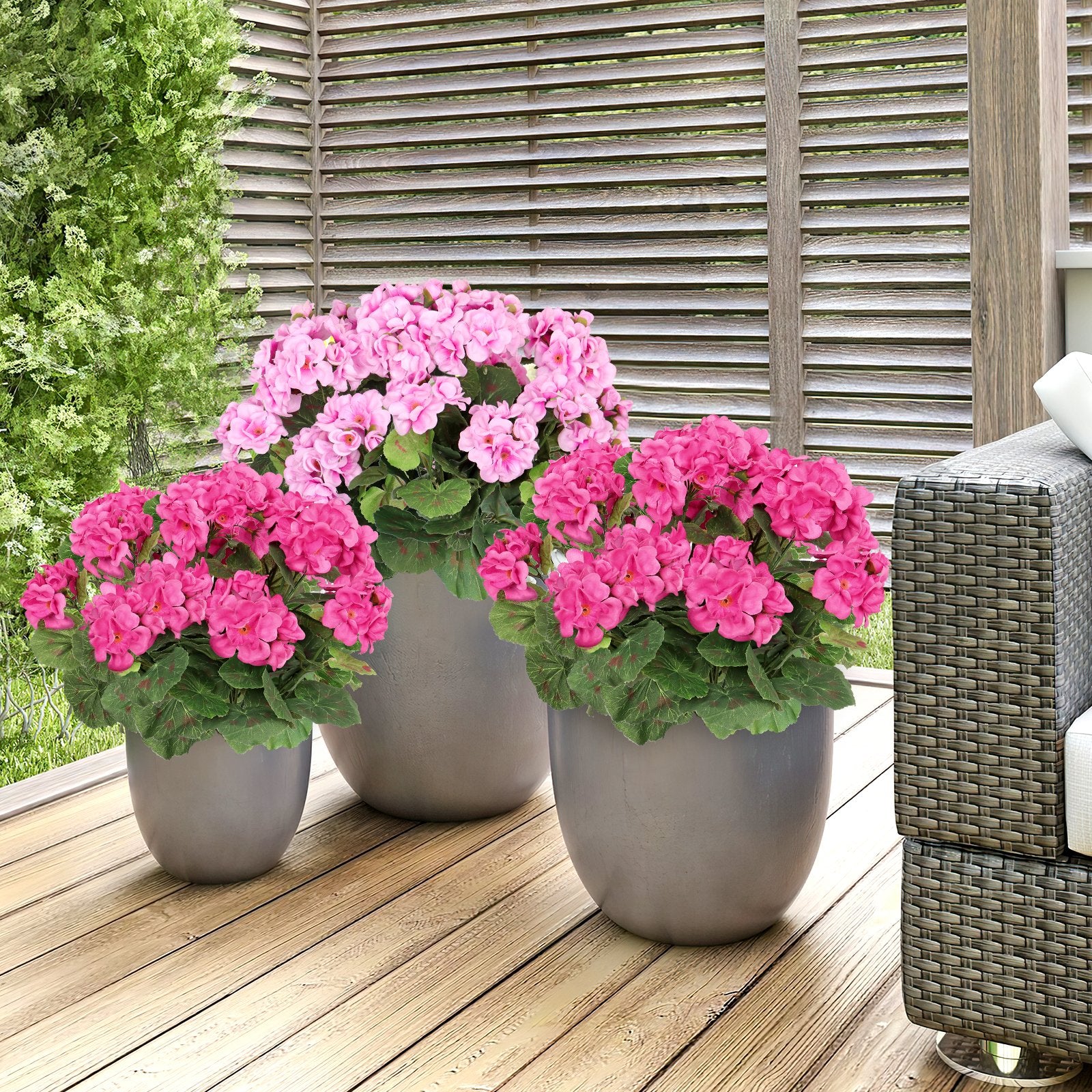 Three potted artificial geranium flowers in pink and rose red on a wooden deck with a slatted wooden fence and outdoor furniture.