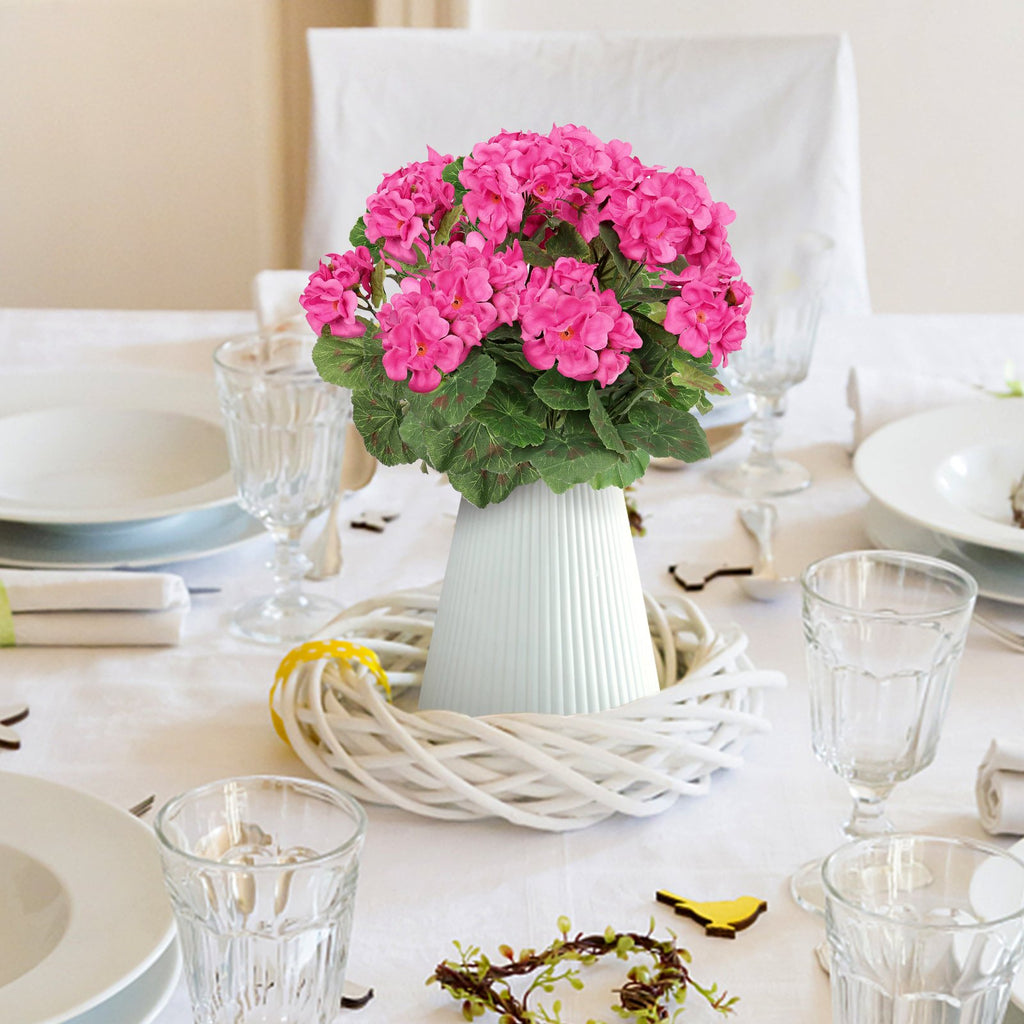 Artificial pink geranium flowers in a white vase on a table setting with plates and glasses