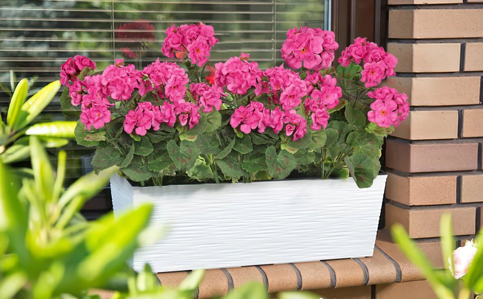 Two bundles of artificial rose red geranium silk flowers in a white planter box on a windowsill.