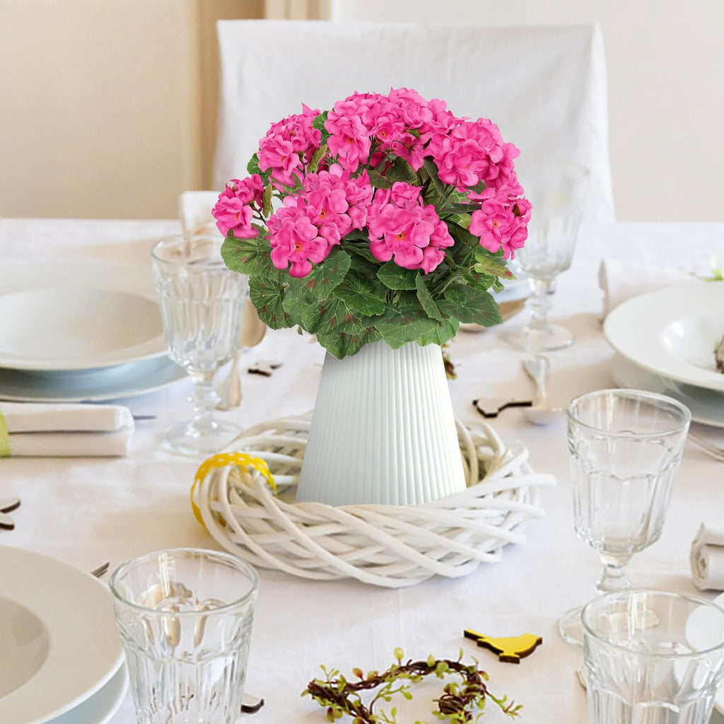 Table setting with a vase of pink artificial geranium flowers as a centerpiece, surrounded by a white wreath.