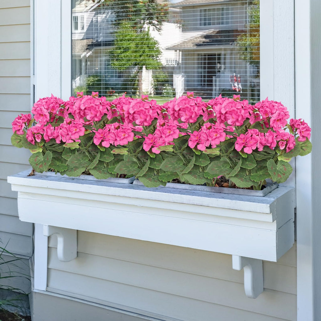 Two bundles of artificial pink geranium flowers in a white window box, with a house reflected in the window behind.