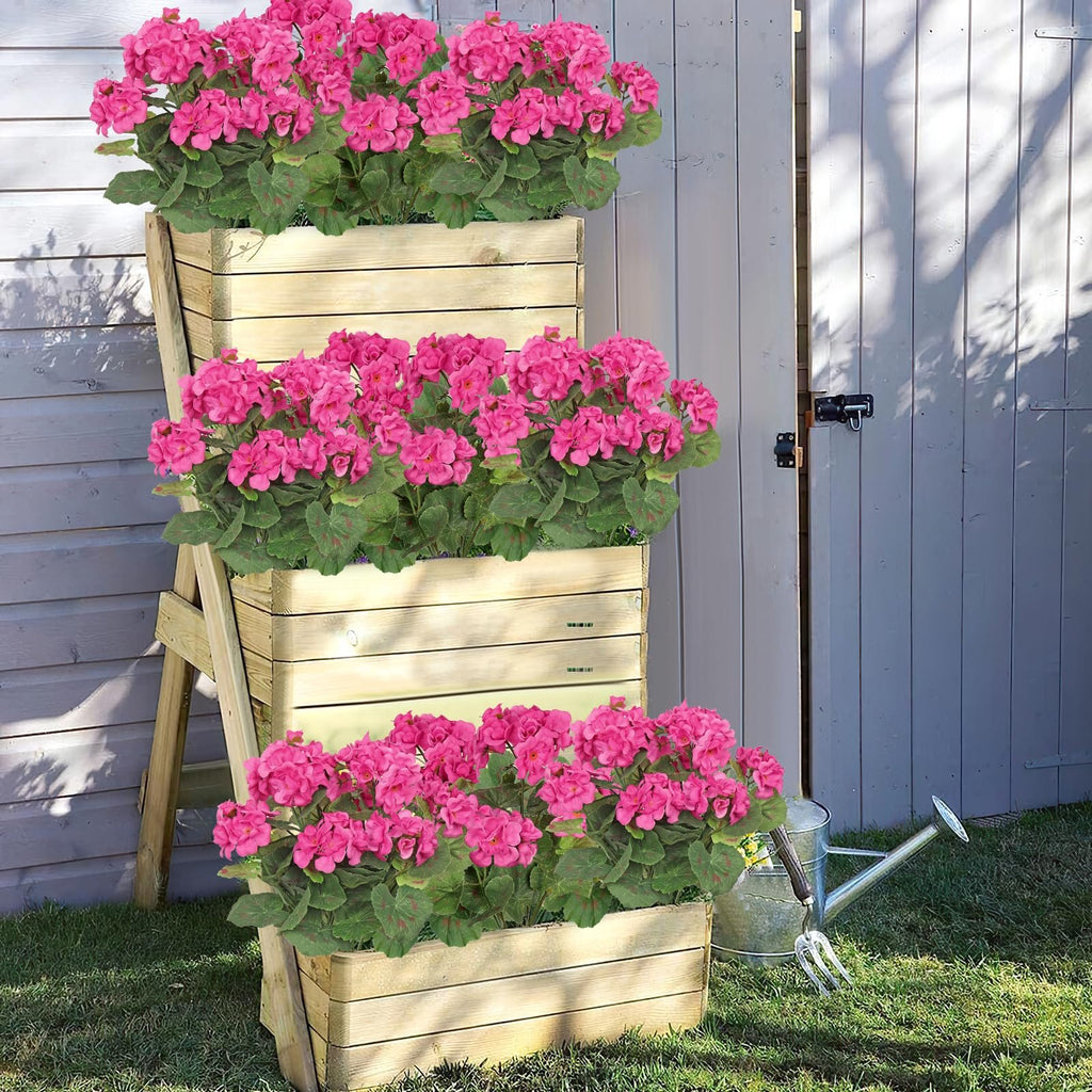 Three tiered wooden planter boxes filled with vibrant pink artificial geranium flowers, set on a grassy lawn next to a gray wooden door.