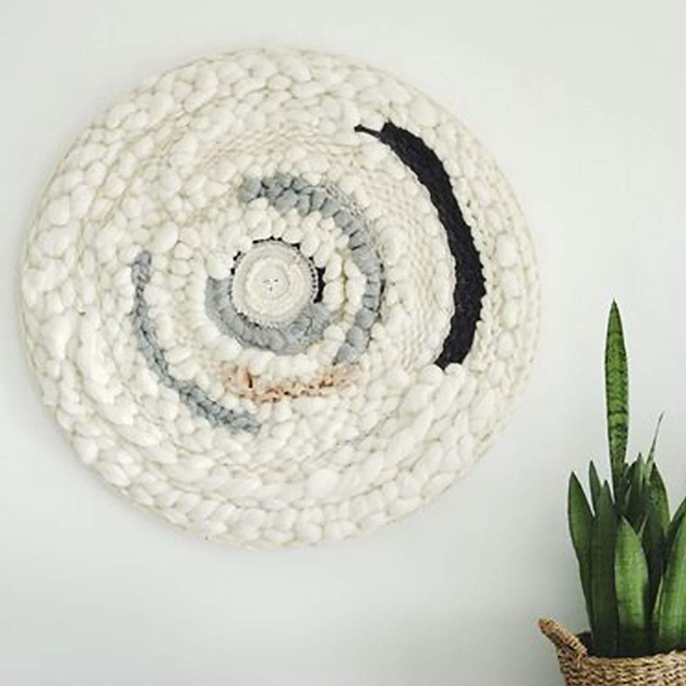 Round woven wall hanging with white, grey, and black yarn in a spiral pattern, displayed on a white wall next to a snake plant.