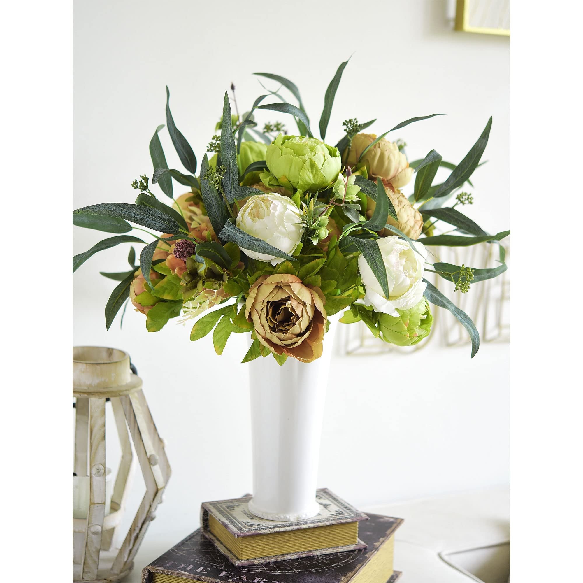 Artificial peony bouquet in a white vase on stacked books, with a rustic lantern to the left.