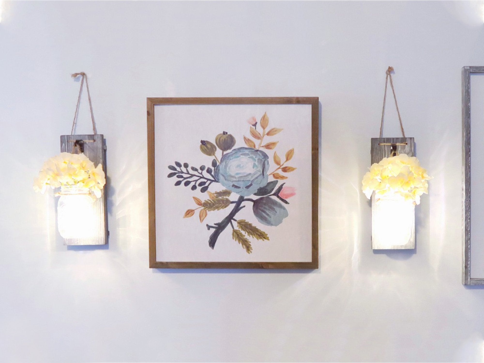 Two rustic wood wall sconces with mason jars and LED lights flank a framed floral art print on a white wall.
