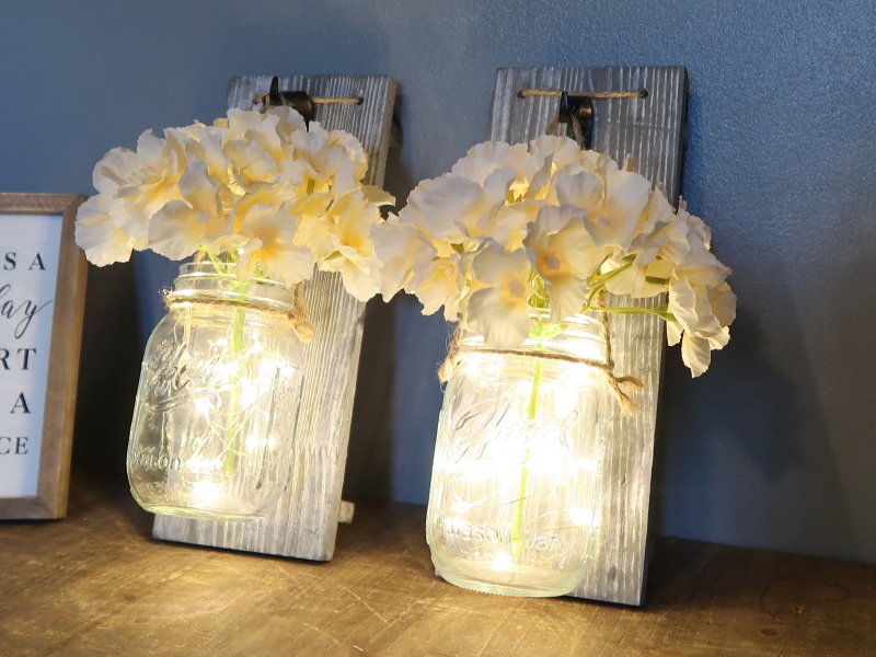 Two rustic wood wall sconces with mason jars filled with white flowers and LED string lights, displayed on a shelf.