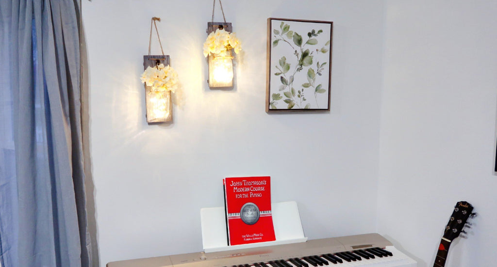 Two rustic wood wall sconces with mason jars and faux flowers hang on a wall next to a framed botanical print and a digital piano.