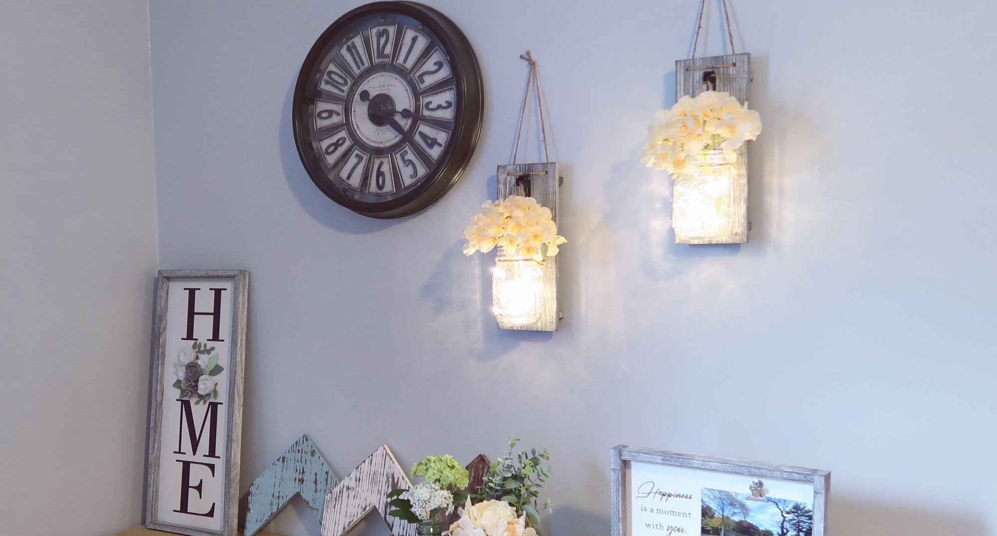 Two rustic wood wall sconces with mason jars and faux flowers hang on a wall next to a large clock.