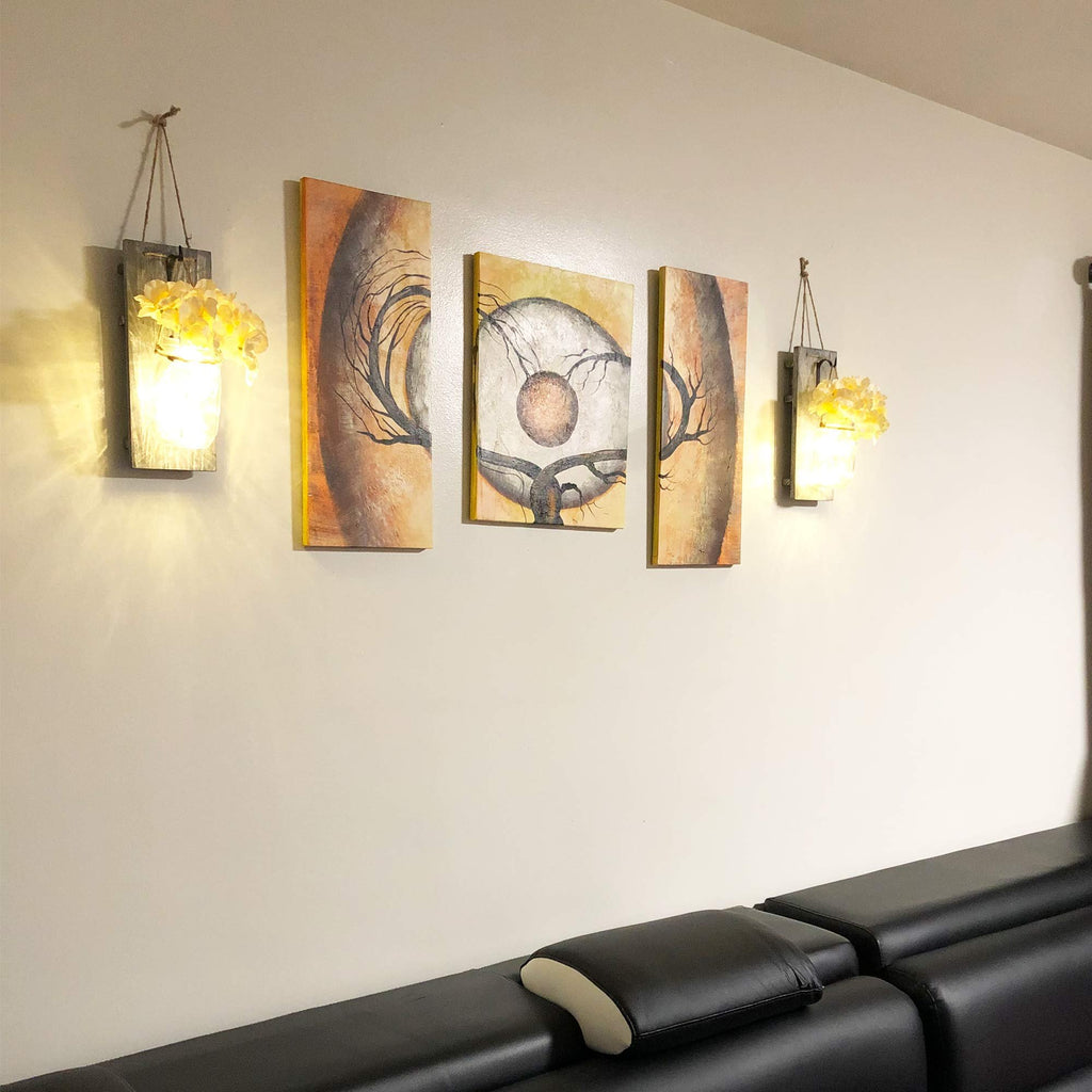 Two rustic wood wall sconces with mason jars and yellow flowers hang on a wall next to a triptych painting above a black leather sofa.