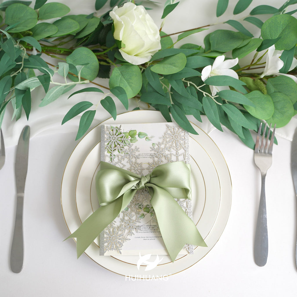 Wedding invitation with sage green ribbon and snowflake design on a plate with cutlery and greenery