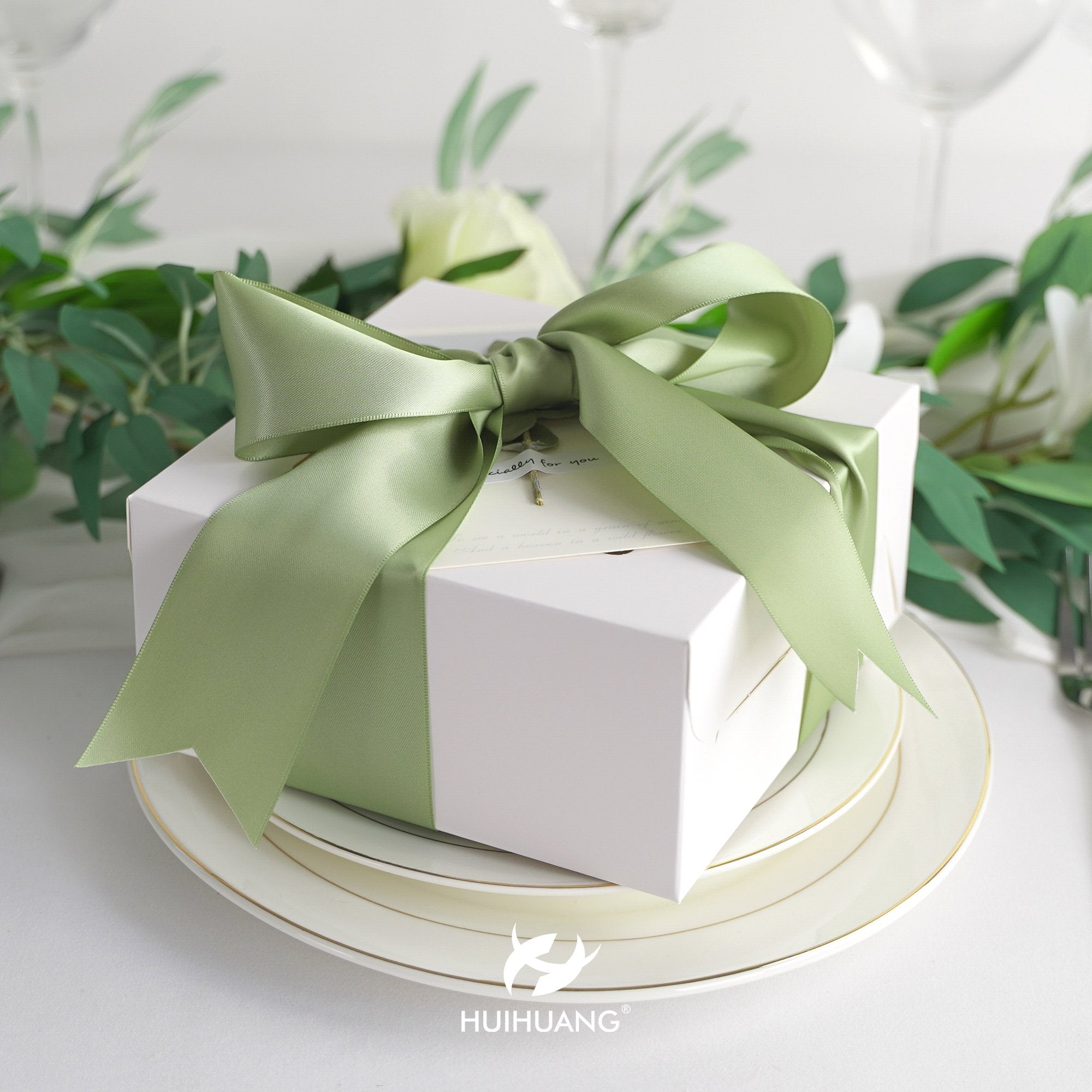 White gift box with a sage green satin ribbon bow, set on plates with greenery and glassware.