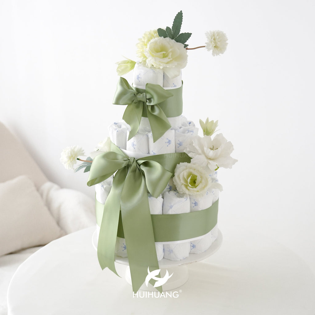 Three-tier diaper cake decorated with sage green ribbon and white flowers, on a white cake stand.
