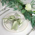 Sage green ribbon tied in a bow on a place setting with a decorative card and greenery