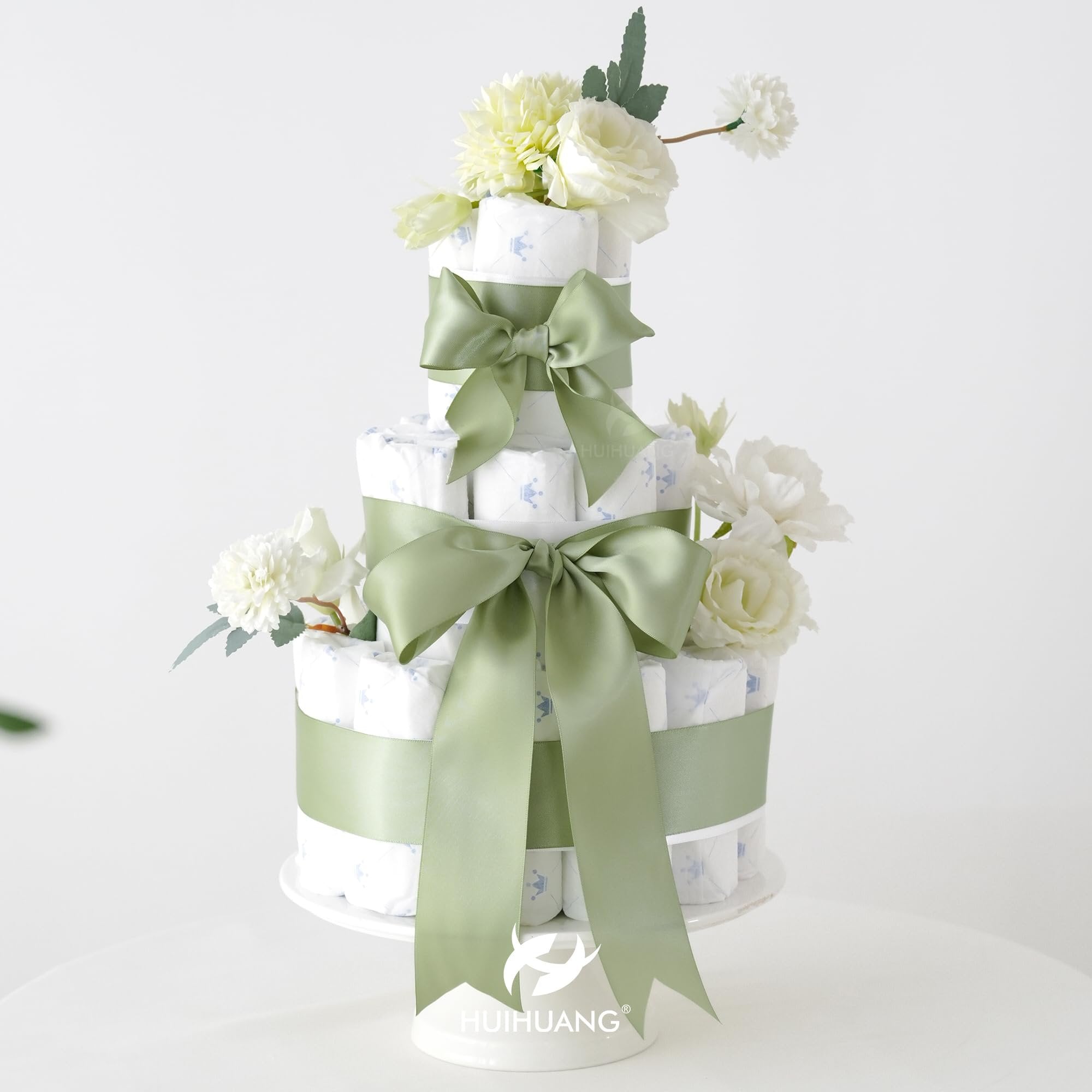 Three-tier diaper cake decorated with sage green ribbon and white flowers, on a white cake stand.