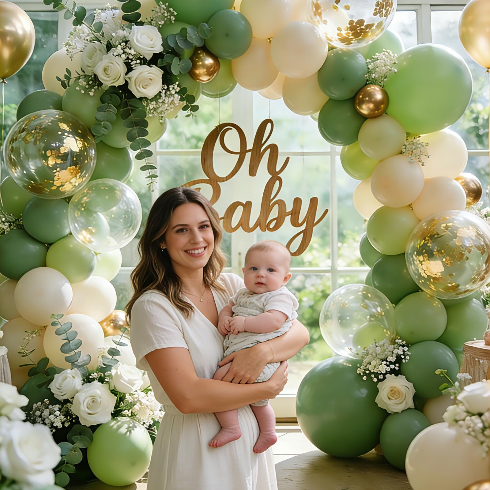 Woman holding baby in front of sage green and gold balloon arch with 'Oh Baby' sign