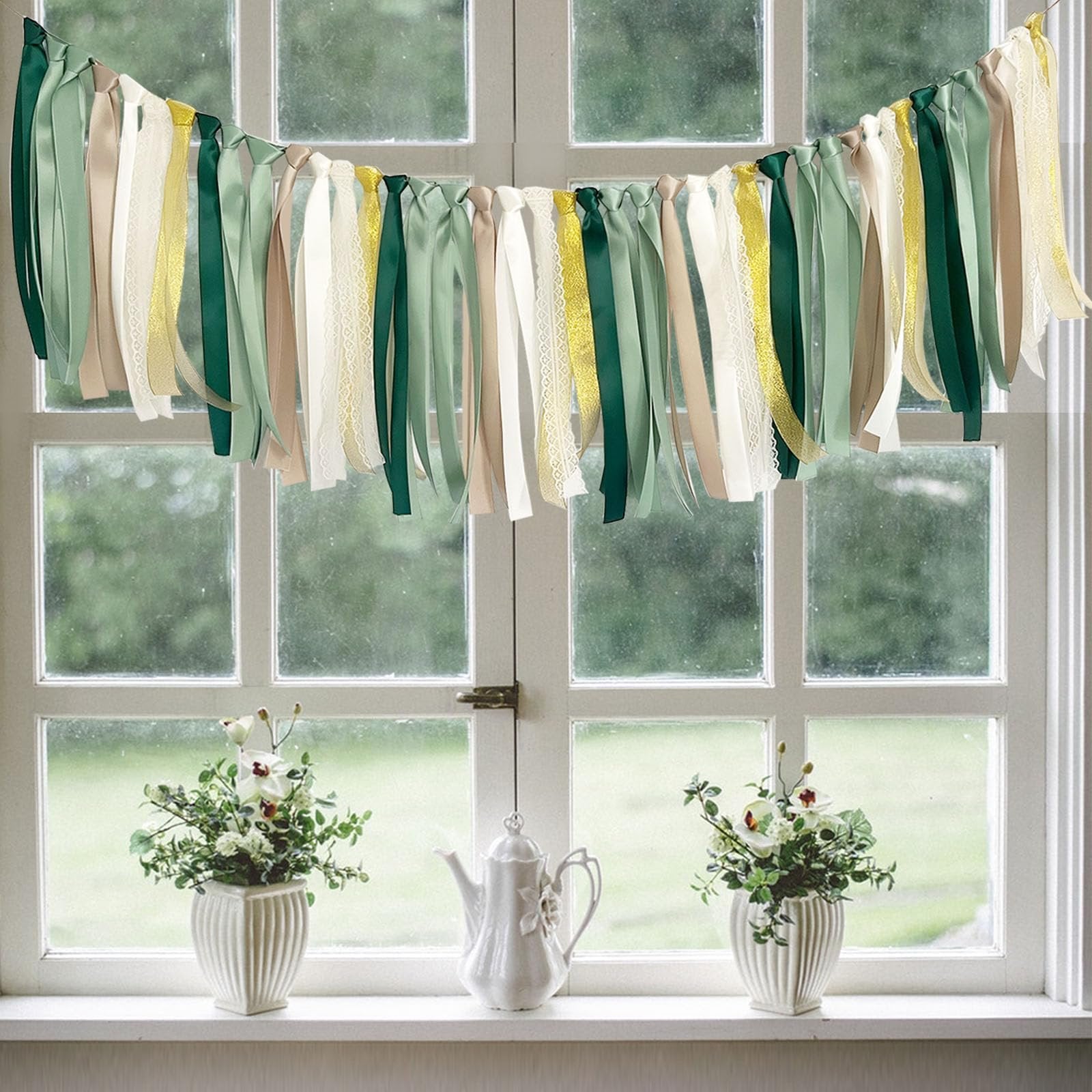 Sage green ribbon garland with cream and gold accents hanging across a window, with decorative vases and a teapot on the windowsill below.