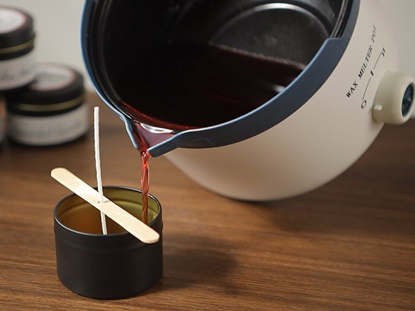 Pouring melted wax from a pot into a candle jar with a wick and wooden stabilizer.