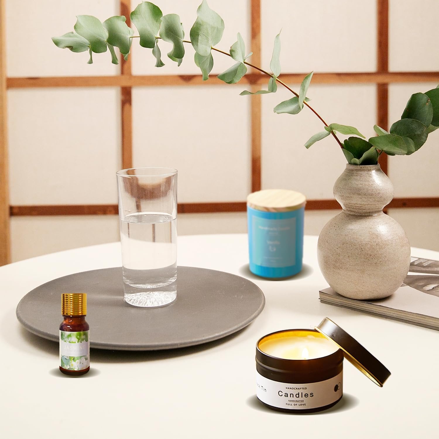 Scented candle in a tin with lid open, next to a glass of water, essential oil bottle, and decorative vase with eucalyptus.
