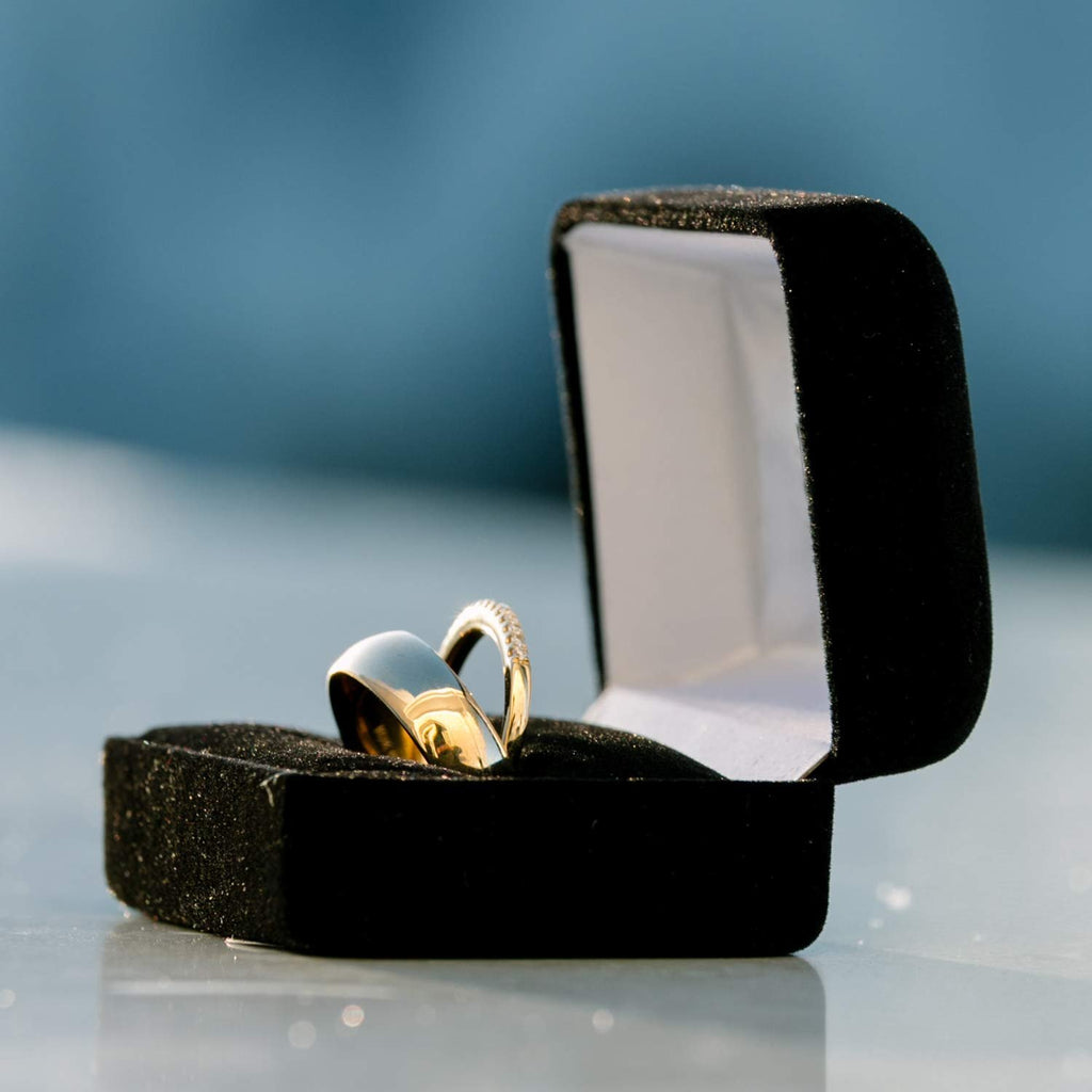 Two gold wedding rings in an open black velvet ring box against a blurred blue background.