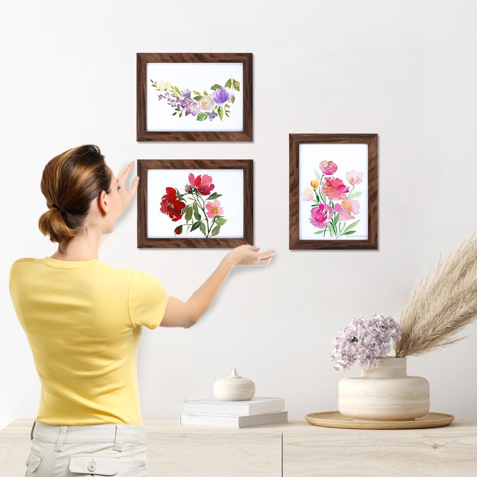 Woman hanging rustic brown wood picture frames with floral art on white wall