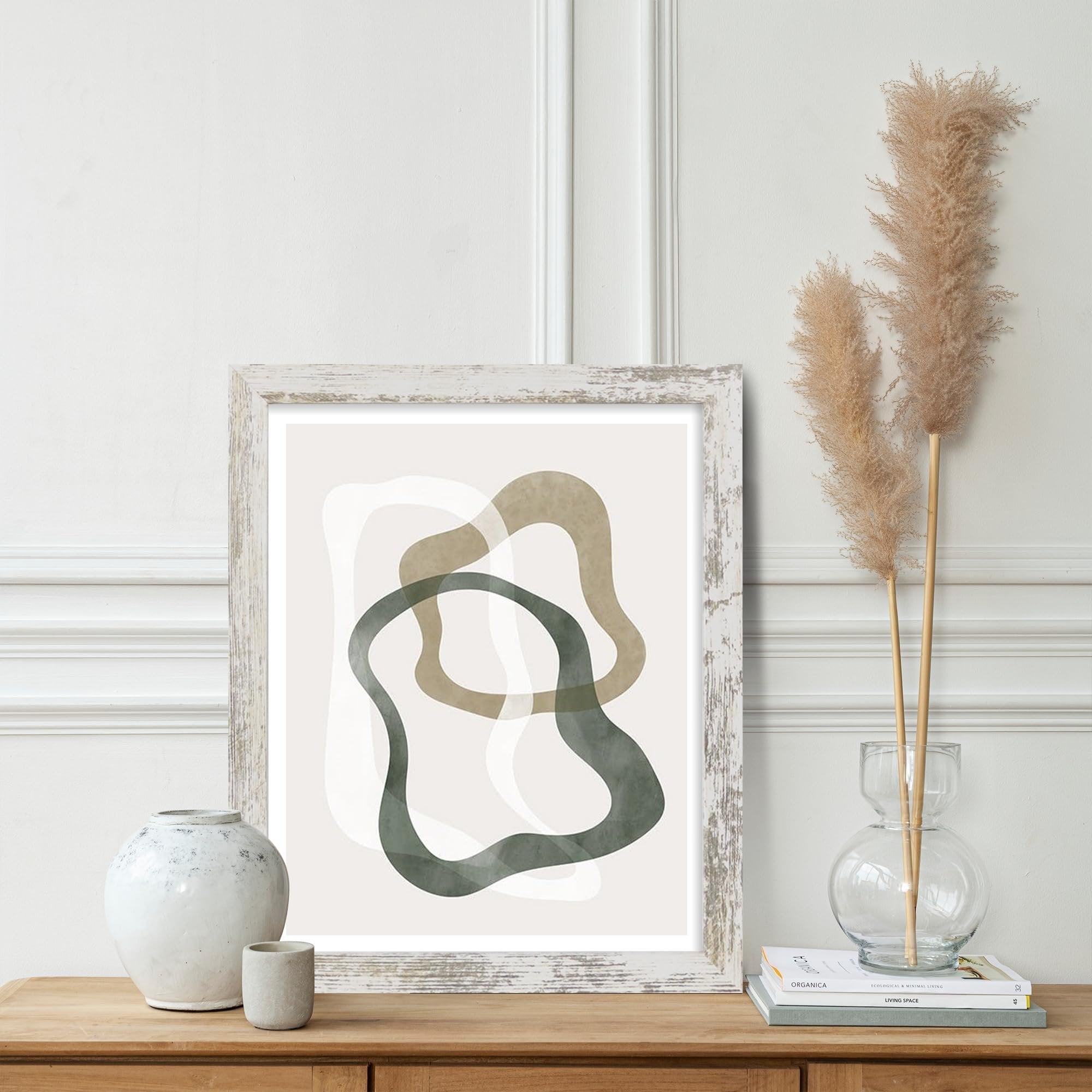 Distressed white wood picture frame displaying abstract art on a wooden table with decorative vases and books.