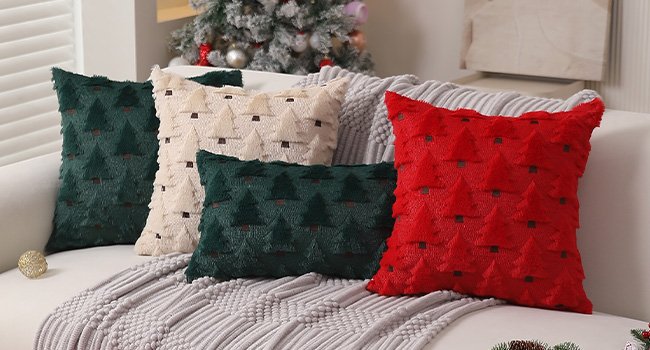 Four Christmas tree patterned pillow covers in red, beige, and green arranged on a sofa with a knitted throw.
